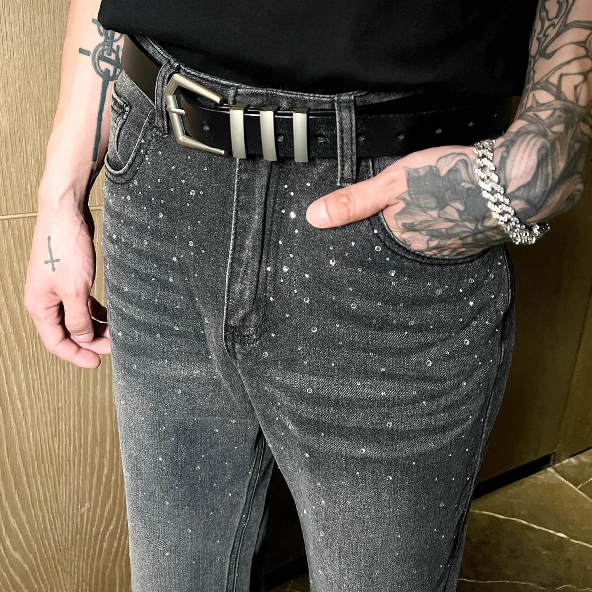 メンズのグレーウォッシュデニムパンツ。全体にクリスタル装飾が散在し、黒レザーベルトに金属バックル付き。