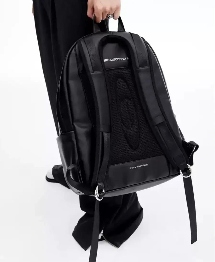 メンズの黒レザー調のバックパックの背面。メッシュのバックパネルと太いストラップ、金属リング付きの長い黒ベルト、上部のハンドルが写る。
