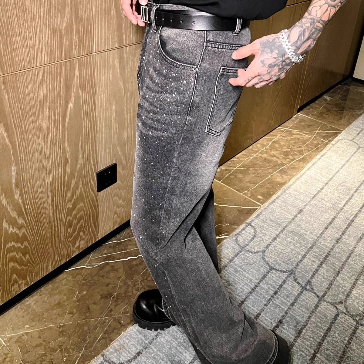 メンズのグレーデニムパンツ。左脚全体にクリスタル装飾が散在し、ホワイトステッチ、黒ベルト、ワイドシルエット。
