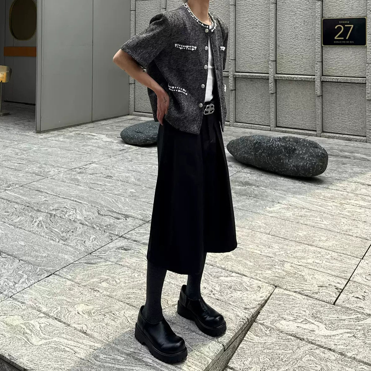 メンズの片手を腰に当てた着用の全身。パール付き黒ツイードジャケットに、ひざ丈の黒ワイドパンツと厚底の黒い靴
