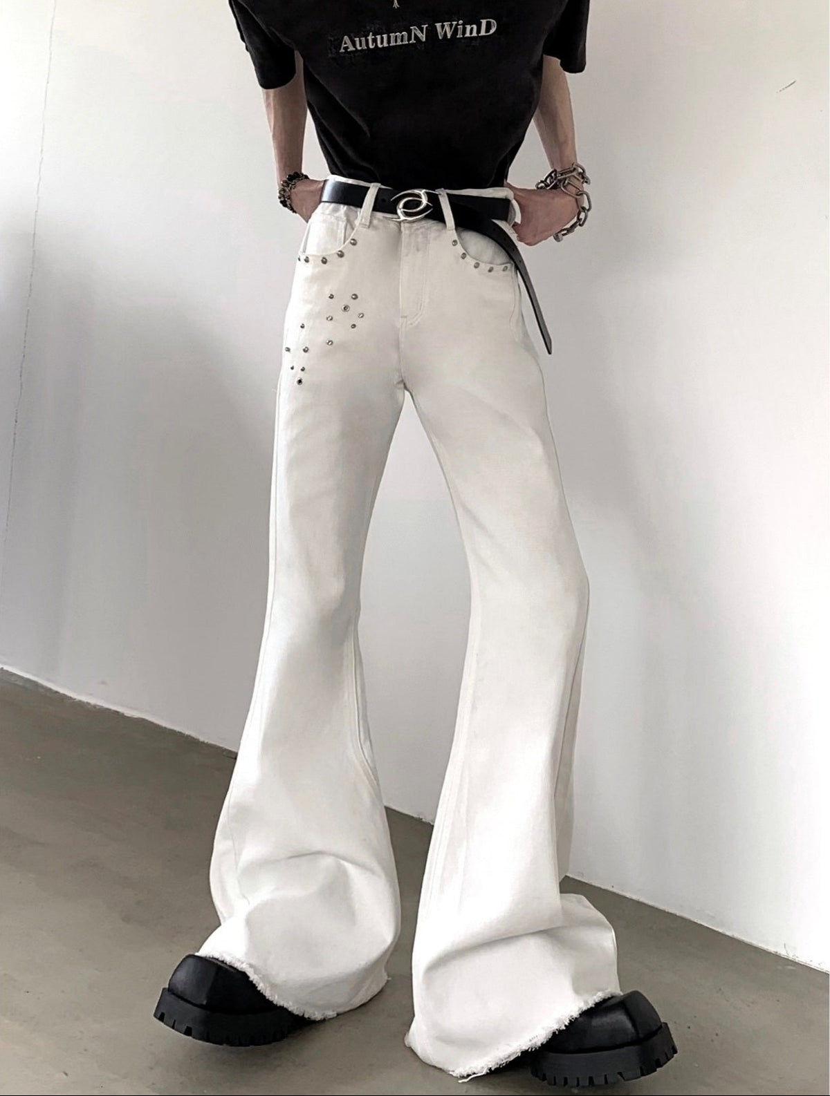 メンズの白デニムのフレアパンツ。腰部分にスタッズ装飾が散らし配置され、すそはドレープを伴ったワイドシルエット。黒ベルト着用。