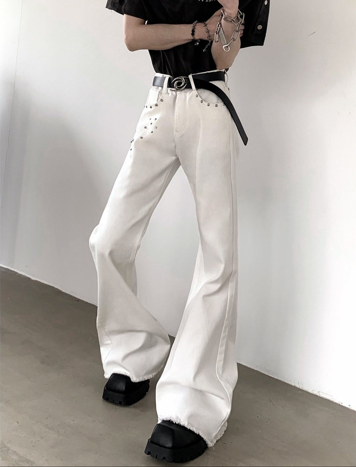 メンズの白デニムパンツ。ハイウエスト、フレアシルエット。ウエスト周りに銀色スタッズが配置された装飾あり。黒ベルト付き。裾はフリンジ加工。