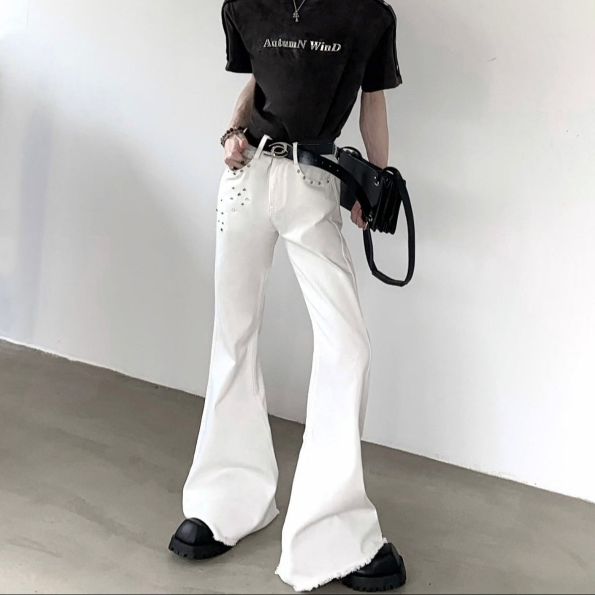 メンズの白デニムのフレアパンツ。ウエスト部分にスタッズ装飾が施され、脚部分は大きくドレープする流れるようなシルエット。裾は広がり気味で素足を見せる着丈。