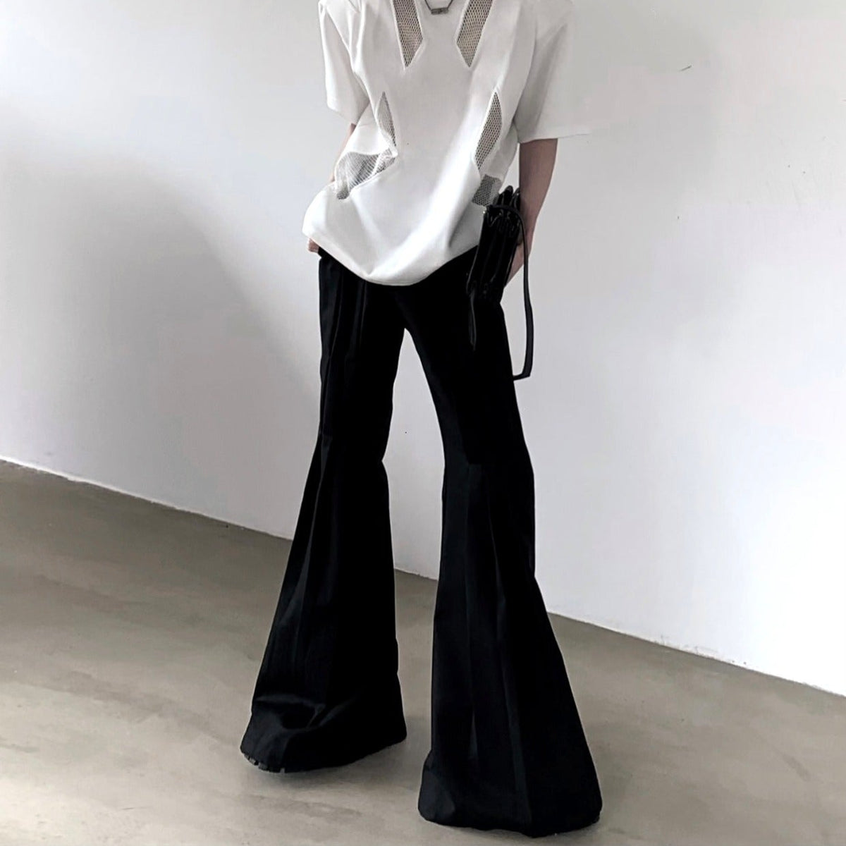 メンズの白いメッシュ切替Tシャツに黒のフレアスラックスを合わせた全身。裾が大きく広がり、小さな黒バッグを手に持つ。