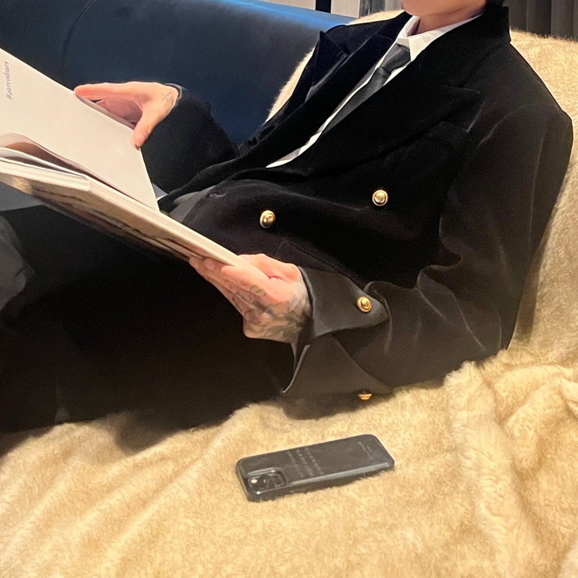 メンズのブランケットの上で雑誌を読む着用。黒のベロア調ジャケットは金ボタンと折り返しカフスが見え、白シャツとネクタイを合わせる。