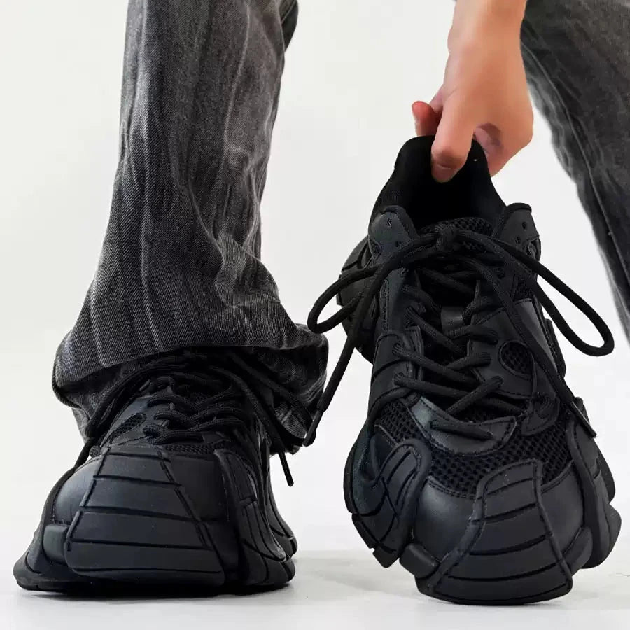 黒いレザーとメッシュ素材のスニーカー。厚底デザインで、黒い紐靴。グレーのテキスタイルパンツを合わせた足元の着用イメージ。