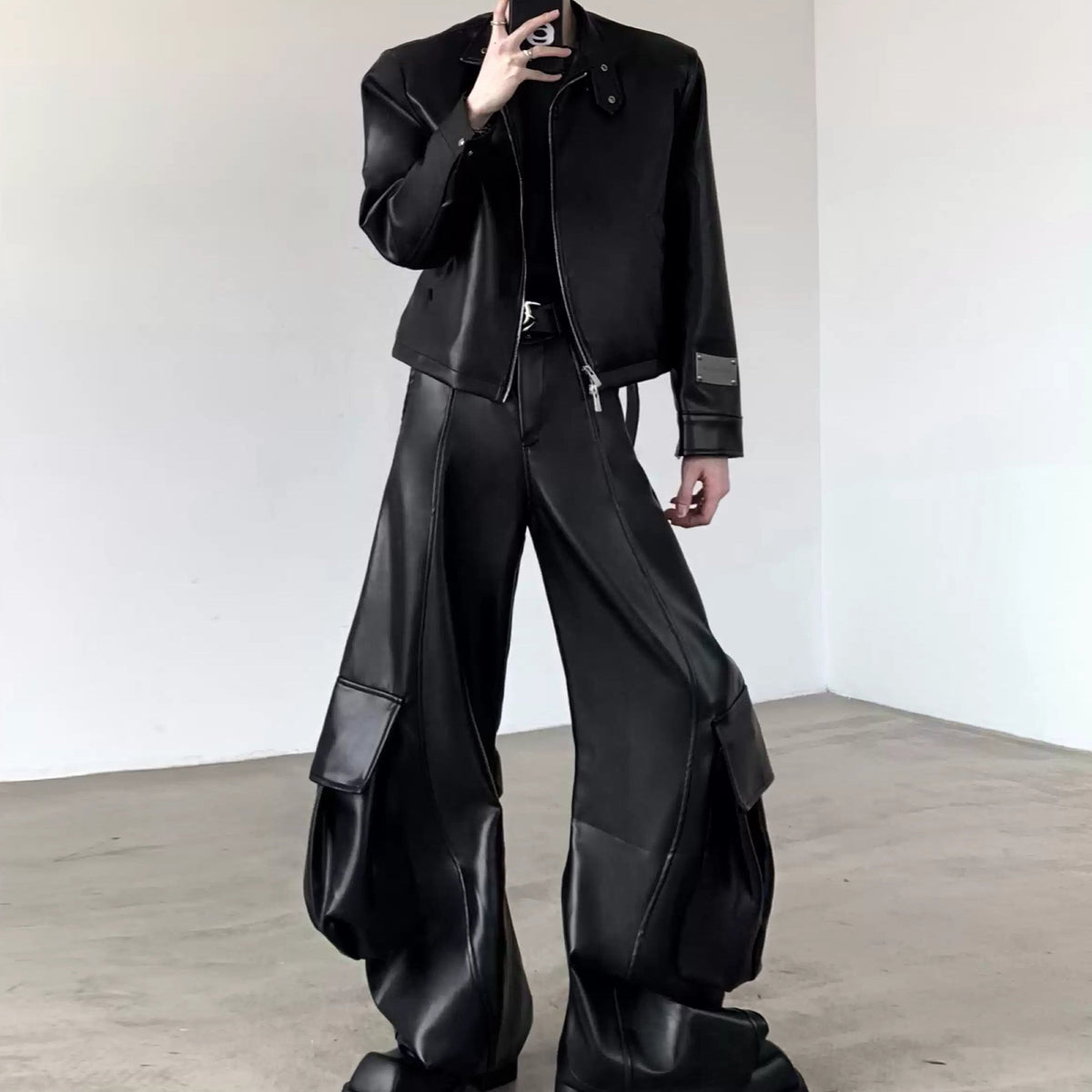 メンズの黒レザーのショートジャケット。ノーカラー、ボタン留め、袖口にレザー切替。ウエストゆったり、バルーン裾の黒レザーパンツと合わせたセットアップスタイル。