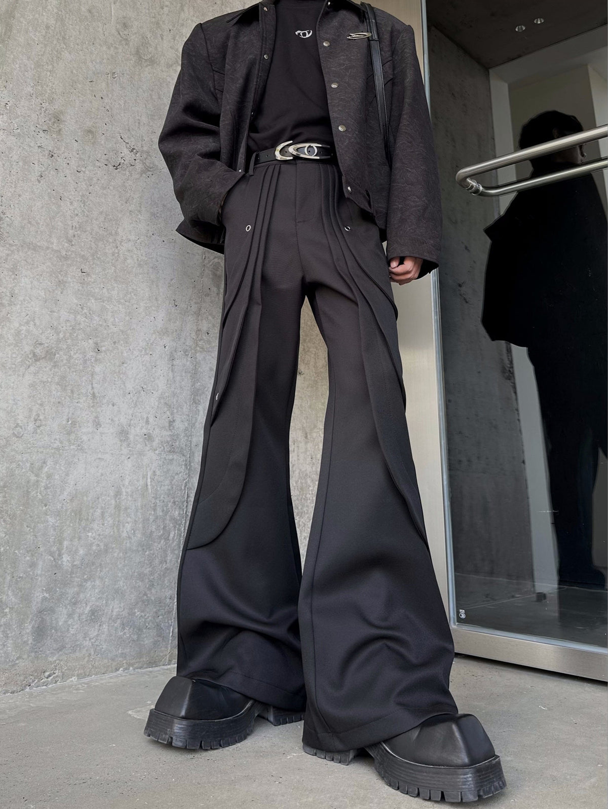 メンズの黒のフレアパンツを低い位置から見た正面。腰の重ねプリーツとハトメ金具があり、裾が大きく広がる。