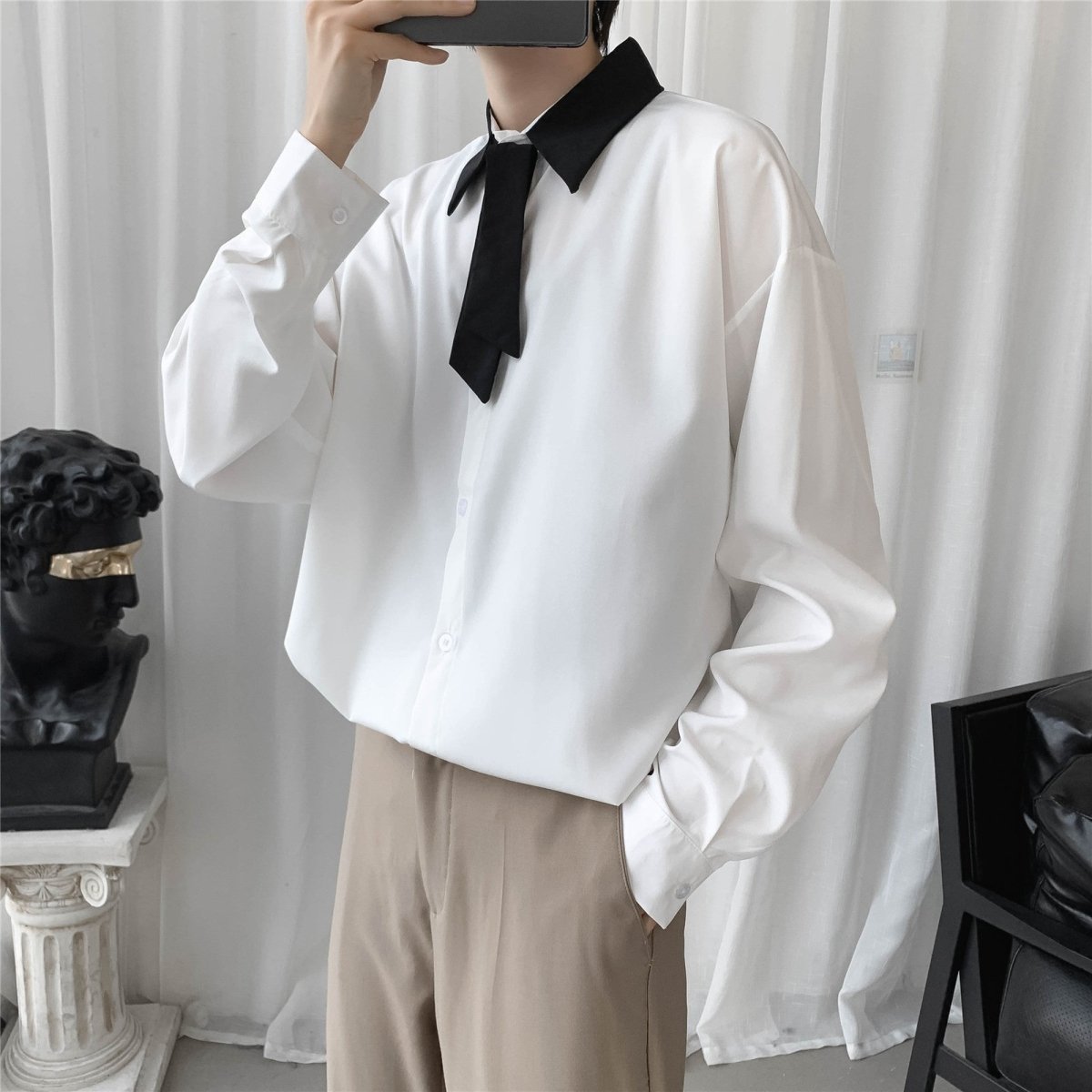 白のオーバーサイズシャツ。黒い衿と黒いリボンタイが付き、前開きボタン式。長袖でゆったりしたシルエット。