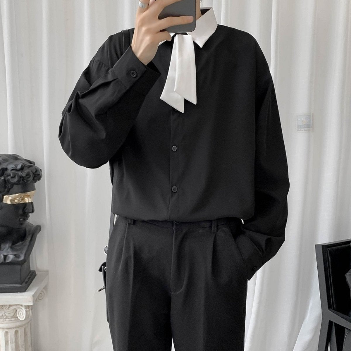 黒のオーバーサイズシャツ。白いリボンタイを首元にあしらい、前ボタン仕様で、落ち感のある素材。黒スラックスと合わせたモノトーン装い。