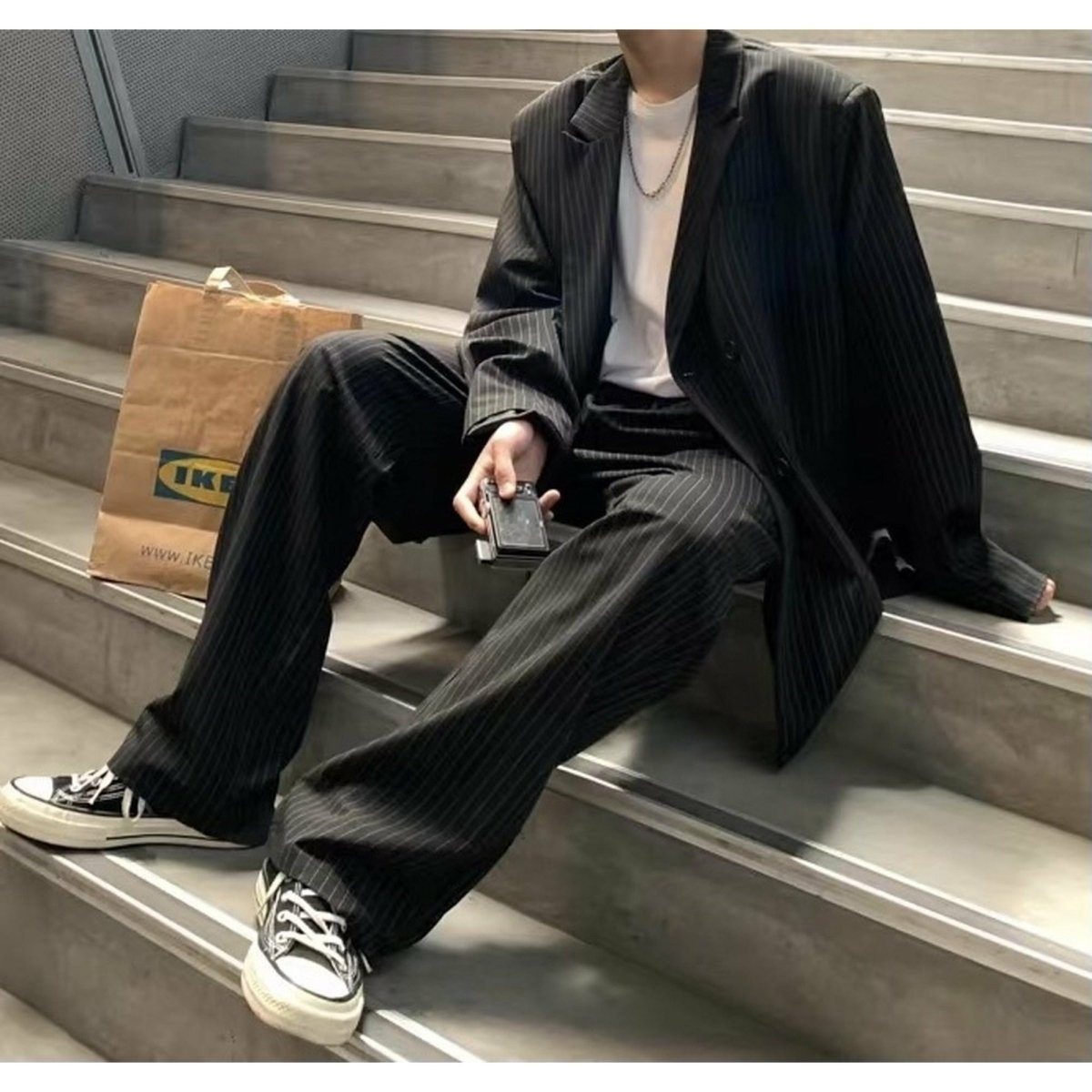 黒のピンストライプセットアップ。オーバーサイズのシングルジャケットと同柄のワイドパンツで構成、ノーカラー襟で開いた前合わせ。
