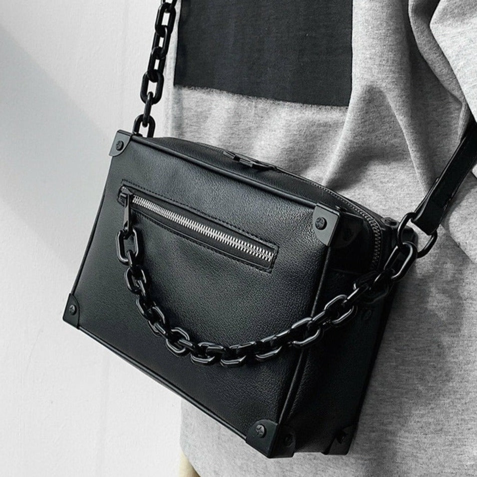 黒レザーのスクエアショルダーバッグ。フロントにジッパーポケット、太めのチェーンストラップと四隅の金具装飾が付いた構造。