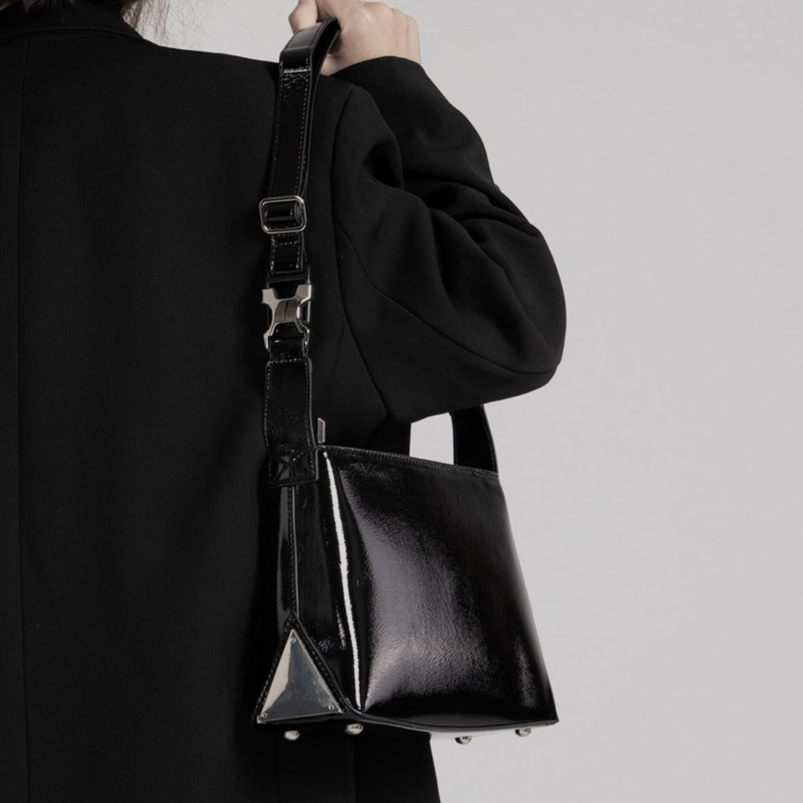 メンズの黒エナメルレザーのショルダーバッグ。四角いシルエットに三角形の金属プレートが装飾され、調節可能な黒レザーストラップと金属バックル付き。