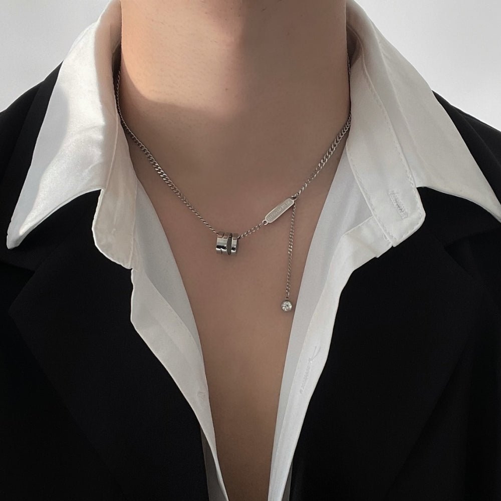 シルバーチェーンネックレス。クリスタルペンダントと金具装飾が付き、黒ジャケットと白シャツの上から着用されている